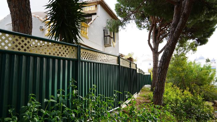 A long green steel fence with a cream trellis top, between pine trees